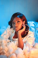 A woman with intricate makeup featuring small decorative elements around her eyes, wearing large hoop earrings. Her hand is gracefully resting on her face. The lighting casts a mix of blue and warm tones, creating a soft, dreamy atmosphere. She is surrounded by fluffy, cloud-like material, adding to the surreal effect.