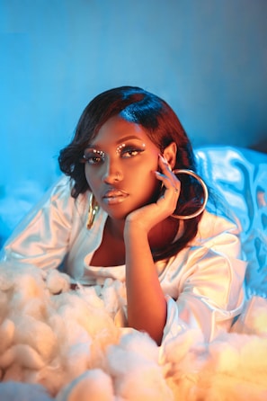 A woman with intricate makeup featuring small decorative elements around her eyes, wearing large hoop earrings. Her hand is gracefully resting on her face. The lighting casts a mix of blue and warm tones, creating a soft, dreamy atmosphere. She is surrounded by fluffy, cloud-like material, adding to the surreal effect.