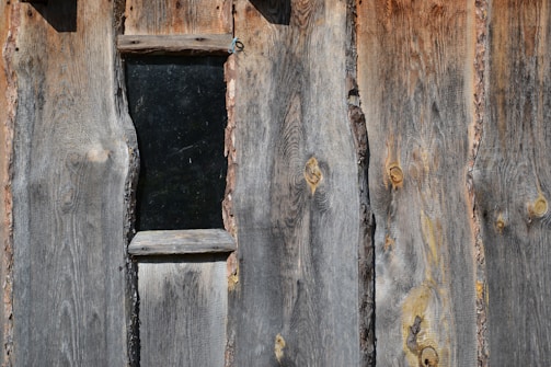 Rough wooden planks with visible grain patterns and knots are arranged vertically. A small rectangular window is embedded within the planks, bordered by a raw wood frame. The wood exhibits weathered textures and variations in tone, from gray to brown.