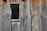 Rough wooden planks with visible grain patterns and knots are arranged vertically. A small rectangular window is embedded within the planks, bordered by a raw wood frame. The wood exhibits weathered textures and variations in tone, from gray to brown.
