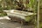 An old, rusted bathtub is situated outdoors on a stone pathway surrounded by lush green foliage and trees. Sunlight filters through the leaves, casting dappled shadows on the bathtub and the ground.