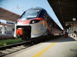 A modern train with a sleek front design is parked at a railway station platform. The train features a silver body with blue and orange accents, and there are multiple people on the platform, some with luggage, suggesting they are either boarding or waiting.