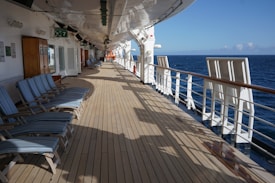 A wooden deck on a cruise ship with several striped lounge chairs arranged along the left side. On the right, a railing overlooks a vast expanse of blue ocean. The deck is mostly empty, with life preservers and small equipment visible in the distance. The sky is clear and sunny, contributing to a serene and tranquil atmosphere.