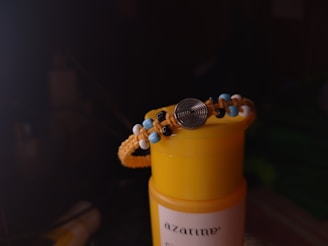 A bracelet with colorful beads, including blue, black, and white, is draped over the top of a yellow cylindrical container. The bracelet features a central metallic circular element with spiral grooves. The background is dark, creating a contrast with the bright foreground.