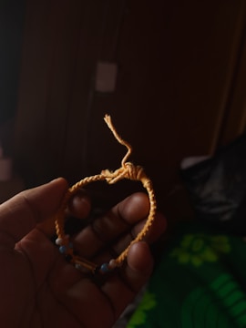 A hand is holding a handmade bracelet with a woven design, made primarily of yellow cord. It includes small beads in varying colors such as blue and black. The background is dark, emphasizing the bracelet.