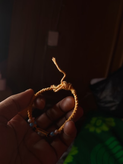 A hand is holding a handmade bracelet with a woven design, made primarily of yellow cord. It includes small beads in varying colors such as blue and black. The background is dark, emphasizing the bracelet.