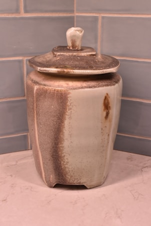 A ceramic jar with a lid, featuring a subtle gradient from light beige to brown tones. It is placed against a tiled wall with a light gray and blue hue and rests on a marble or stone surface. The finish of the jar has a slightly rustic appearance with speckled texture.