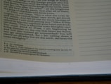 A close-up view of an open book showing text on both left and right pages. The left page contains printed text in a paragraph format with some footnotes at the bottom. The right page is mostly empty with lined spaces for writing notes.