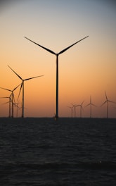 Several wind turbines are situated offshore, their large blades silhouetted against a gradient sky transitioning from orange to blue as the sun sets. The sea is calm and dark in the foreground, adding contrast to the serene landscape.