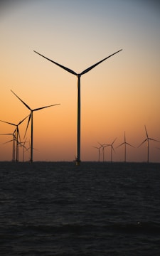 Several wind turbines are situated offshore, their large blades silhouetted against a gradient sky transitioning from orange to blue as the sun sets. The sea is calm and dark in the foreground, adding contrast to the serene landscape.