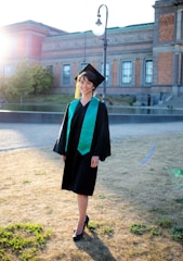 A graduate wearing a black cap and gown with a green sash stands on a patch of sunlit grass. The individual is smiling, and the sunlight creates a warm glow around them. In the background, an old brick building and a streetlamp can be seen.