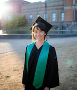 A person in graduation attire, consisting of a black gown and a mortarboard cap with a bright green sash, stands outdoors on a sunny day. The background features a historic building with detailed brick architecture and large windows, and sunlight creates a soft lens flare.