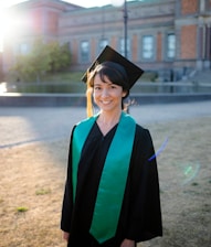 A person in graduation attire, consisting of a black gown and a mortarboard cap with a bright green sash, stands outdoors on a sunny day. The background features a historic building with detailed brick architecture and large windows, and sunlight creates a soft lens flare.