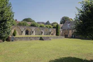 A historical stone structure with arched openings stands at the center of a well-maintained lawn, surrounded by lush green trees and bushes. In the background, residential buildings with sloped roofs are visible, blending seamlessly with the serene environment.