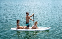 A man is paddleboarding on a calm body of water with two children sitting on the board. The man is standing and paddling while the children are seated in front of him, wearing swimwear. The water appears tranquil and expansive, reflecting the sunlight.