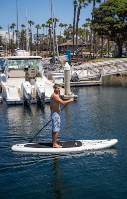 A person is paddleboarding on calm water in a marina. In the background, there are several yachts docked alongside tall palm trees and a clear blue sky.