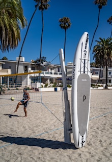 A person is playing volleyball on a sandy beach, with buildings and tall palm trees in the background. A stand-up paddleboard is leaning against a volleyball net post. The sky is clear and blue, giving the scene a vibrant and sunny atmosphere.