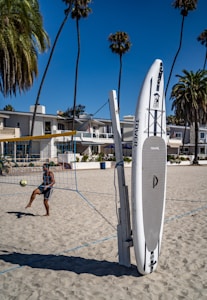 A person is playing volleyball on a sandy beach, with buildings and tall palm trees in the background. A stand-up paddleboard is leaning against a volleyball net post. The sky is clear and blue, giving the scene a vibrant and sunny atmosphere.