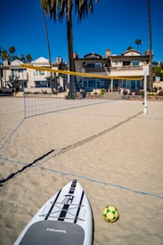 A sandy beach setting with a yellow and blue volleyball net and a nearby paddleboard and a yellow and green ball on the ground. In the background, there are several palm trees and beach houses with architectural features like balconies and multiple windows. The sky is clear and blue, suggesting a sunny day.