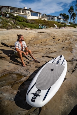 A person with long hair and sunglasses sits on rocky terrain holding a paddle beside a paddleboard. The setting includes a beach with modern houses on the hillside and a clear blue sky. Bicycles and palm trees are visible in the background.
