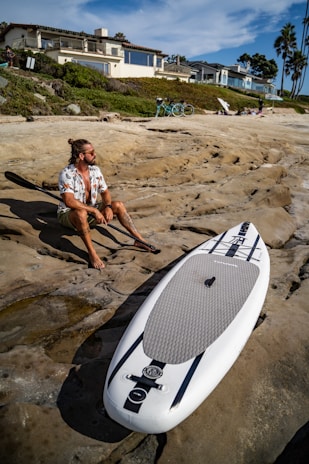 A person with long hair and sunglasses sits on rocky terrain holding a paddle beside a paddleboard. The setting includes a beach with modern houses on the hillside and a clear blue sky. Bicycles and palm trees are visible in the background.