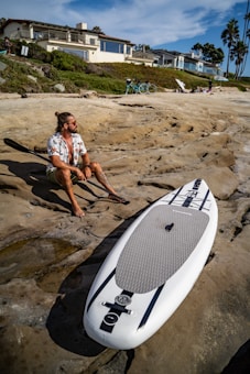 A person with long hair and sunglasses sits on rocky terrain holding a paddle beside a paddleboard. The setting includes a beach with modern houses on the hillside and a clear blue sky. Bicycles and palm trees are visible in the background.