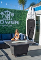 A woman in a bikini and shorts sits on a blue outdoor sofa holding a can of drink. Behind her, there is a large surfboard upright against a wall with the words 'Tower Beach Club.' The background features a green hedge wall with a printed logo of a lifeguard tower and palm trees. A fire pit table is set in front of the sofa, and string lights are visible overhead. The sky is clear and blue, indicating a sunny day.