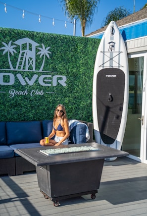 A woman in a bikini and shorts sits on a blue outdoor sofa holding a can of drink. Behind her, there is a large surfboard upright against a wall with the words 'Tower Beach Club.' The background features a green hedge wall with a printed logo of a lifeguard tower and palm trees. A fire pit table is set in front of the sofa, and string lights are visible overhead. The sky is clear and blue, indicating a sunny day.