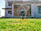 A sprinkler is watering a lush green lawn in front of a modern house with large windows. Water droplets are visibly scattering in the sunlight, creating a refreshing and lively scene.