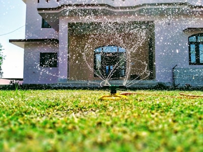A sprinkler is watering a lush green lawn in front of a modern house with large windows. Water droplets are visibly scattering in the sunlight, creating a refreshing and lively scene.