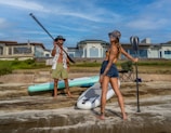 A pair of paddleboarders laughing together while gliding near a sandy beach in Lewes, Delaware.