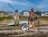 Two individuals holding paddles are on a sandy shore next to stand-up paddleboards. They are dressed casually and appear relaxed. Behind them, modern beachfront houses and a clear blue sky are visible.