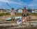 Two individuals holding paddles are on a sandy shore next to stand-up paddleboards. They are dressed casually and appear relaxed. Behind them, modern beachfront houses and a clear blue sky are visible.