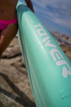A close-up view of a light turquoise inflatable paddleboard held by a person at the beach. The person's hand and hip are visible, wearing pink swimwear. The background reveals rocks and a blurred ocean scene with a bright sky.