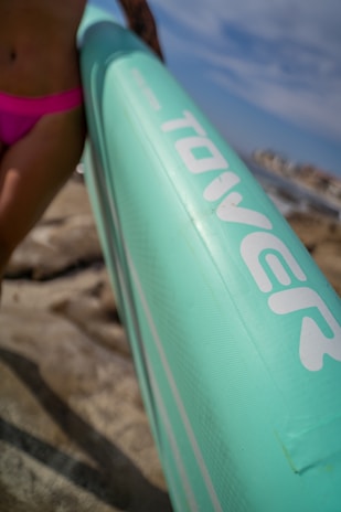 A close-up view of a light turquoise inflatable paddleboard held by a person at the beach. The person's hand and hip are visible, wearing pink swimwear. The background reveals rocks and a blurred ocean scene with a bright sky.