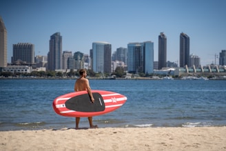 A person holding a red and black paddleboard stands on a sandy beach facing a body of water, with a city skyline featuring tall buildings in the background. The sky is clear and blue.