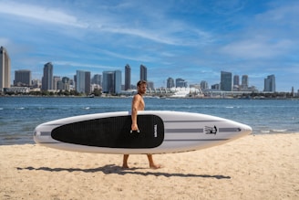 A person is walking along a sandy beach carrying a large paddleboard by their side. In the background, a city skyline with modern high-rise buildings stretches across the horizon. The sky is clear with sparse clouds, and the sea is calm, gently meeting the shore.