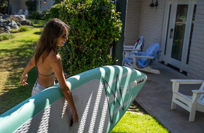 A woman with long hair walks outside in a garden area while carrying a green paddleboard. She is wearing sunglasses and a bikini top. The setting includes greenery, a sunlit lawn, and a patio with white furniture, including a pair of chairs and a door leading into a house.