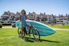 A woman stands on a grassy lawn, holding a bicycle with a large turquoise paddleboard attached to it. She is wearing sunglasses and a blue-striped dress. The background features a row of large, coastal-style houses with pointed roofs under a clear blue sky.