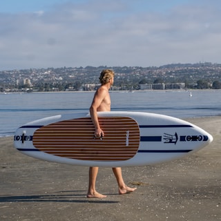 A person with short hair is walking along the shore carrying a paddleboard under one arm. The beach is sandy, and in the background, calm water stretches out with buildings and hills on the horizon under a partly cloudy sky.