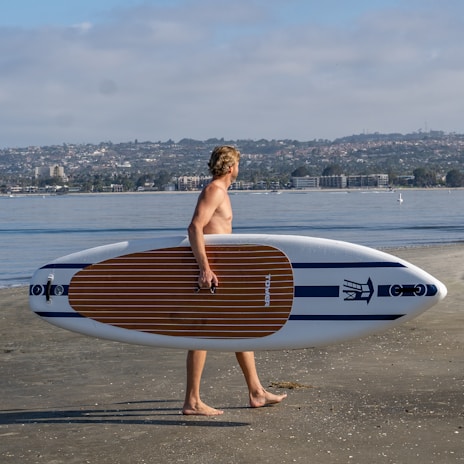 A person with short hair is walking along the shore carrying a paddleboard under one arm. The beach is sandy, and in the background, calm water stretches out with buildings and hills on the horizon under a partly cloudy sky.