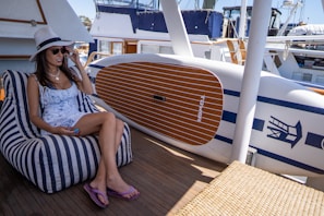 A woman lounging on a stylish deck chair wearing a wide-brimmed hat and an airy kaftan, with sparkling water in the background.