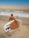 A person carrying a paddleboard walks towards the ocean, with a child standing nearby on the sandy beach. The sky displays a gradient from orange to blue, and waves are gently crashing onto the shore.