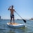 A person is paddling on a stand-up paddleboard in a body of water, wearing floral-patterned shorts. The scene is bright and sunny with a clear blue sky. In the background, a bridge is visible along with some industrial and residential buildings, along with a docked boat.