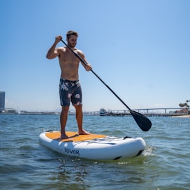 A person is paddling on a stand-up paddleboard in a body of water, wearing floral-patterned shorts. The scene is bright and sunny with a clear blue sky. In the background, a bridge is visible along with some industrial and residential buildings, along with a docked boat.