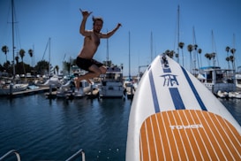 A person is mid-air, jumping off a boat docked in a marina. Nearby, several sailboats and yachts are anchored. A paddleboard branded 'TOWER' with blue and orange stripes is prominently in the foreground. Palm trees line the scene, indicating a sunny and coastal location.