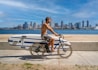 A shirtless man rides a bicycle along a paved pathway adjacent to a sandy beach. The man's bicycle has a surfboard attached to the side. In the background, a body of water separates the pathway from a city skyline with tall buildings under a partly cloudy blue sky.