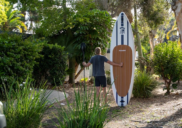 A person is carrying a large paddleboard and standing on a path surrounded by lush green foliage and trees. The paddleboard has a brown and white striped pattern, and the individual is holding a black paddle.