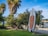 A stand-up paddleboard is positioned against dense greenery, with a sandy beach and body of water visible in the background. Tall palm trees are seen nearby, adding to the tropical feel of the scene. The clear blue sky suggests it is a bright and sunny day.
