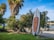 A stand-up paddleboard is positioned against dense greenery, with a sandy beach and body of water visible in the background. Tall palm trees are seen nearby, adding to the tropical feel of the scene. The clear blue sky suggests it is a bright and sunny day.